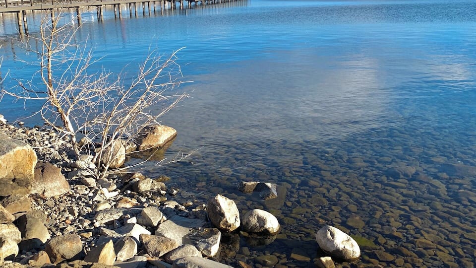 A rocky shoreline with clear water, a wooden pier extending into the lake, and mountains in the background.