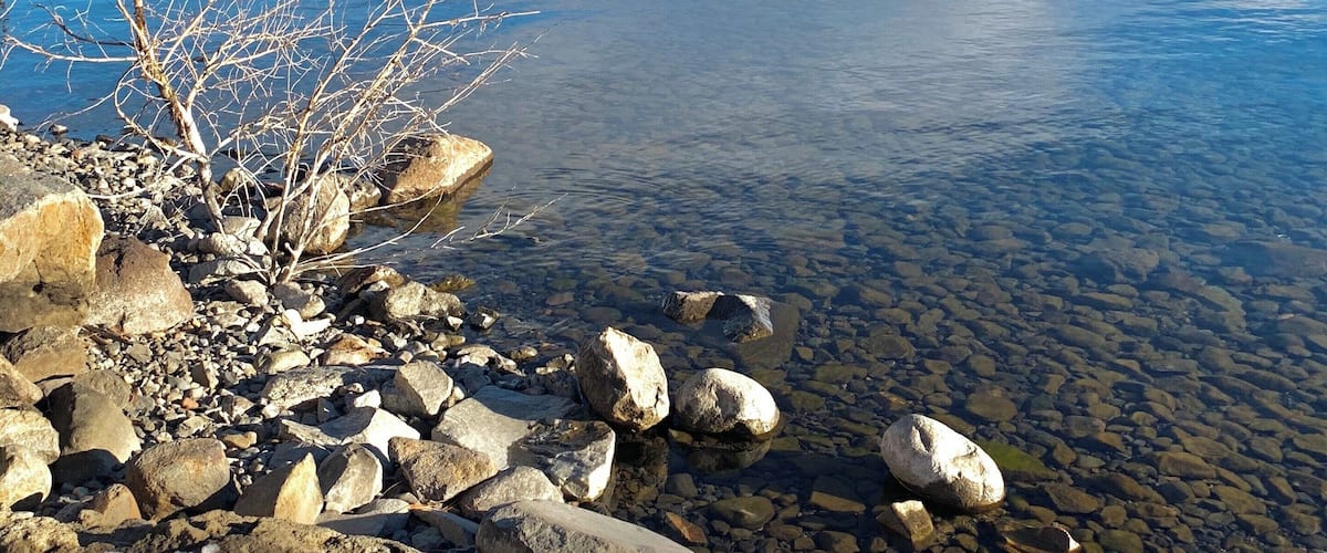 A rocky shoreline with clear water, a wooden pier extending into the lake, and mountains in the background.
