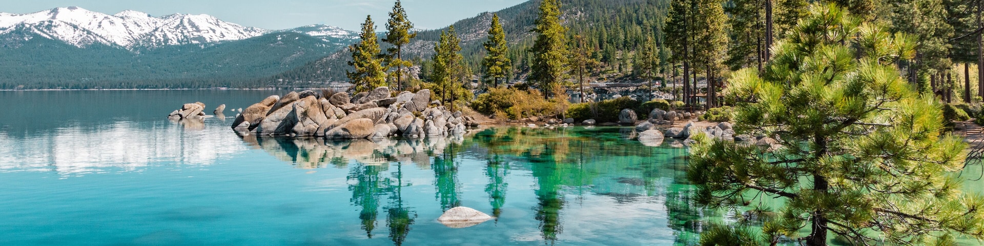 A clear lake with visible rocks and trees, surrounded by mountains and clear skies.