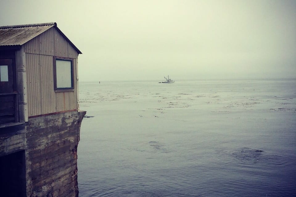 En träbrygga som sträcker sig ut i stilla vatten, med en liten båt i fjärran och en molnig himmel.