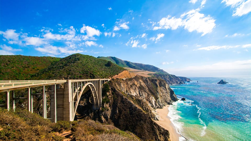 A coastal bridge with a mountainous backdrop and a beach.