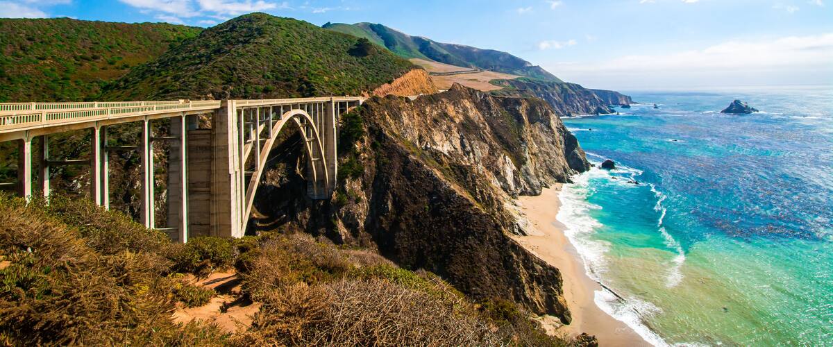 A coastal bridge with a mountainous backdrop and a beach.