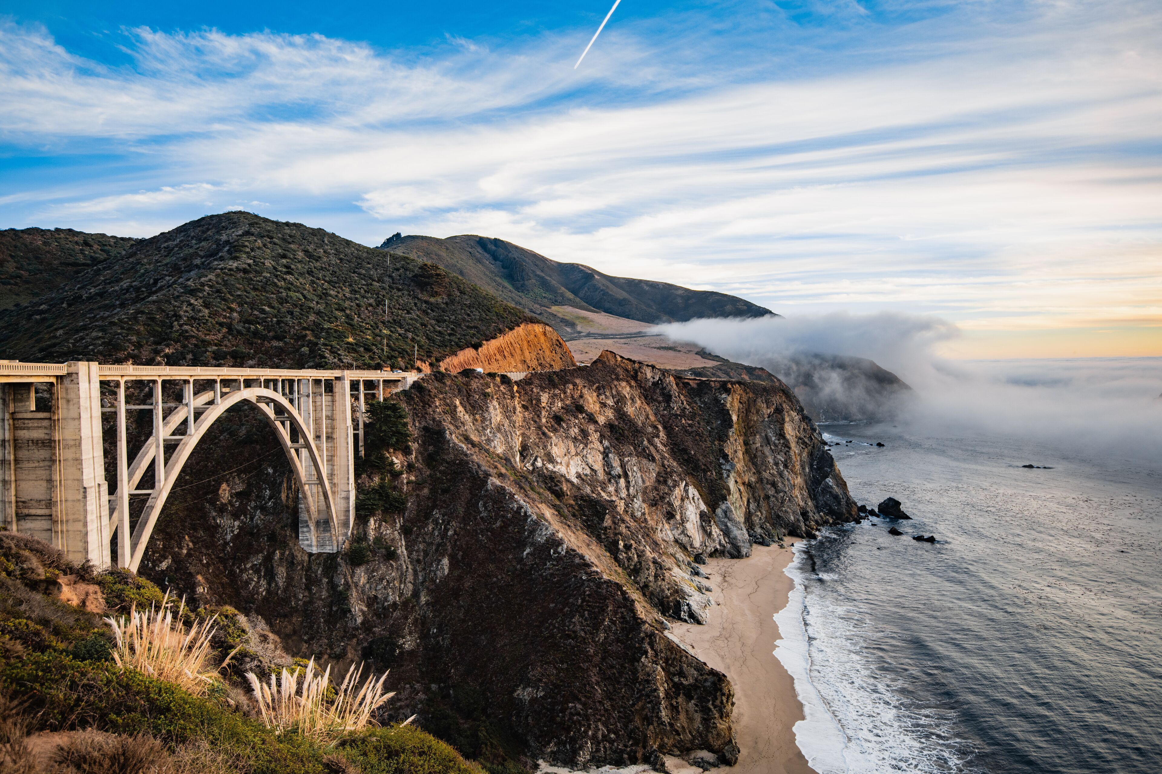 Bixby bridge foggy sunset landscape in Big Sur area on the California coast with ocean waters crashing on the shore.