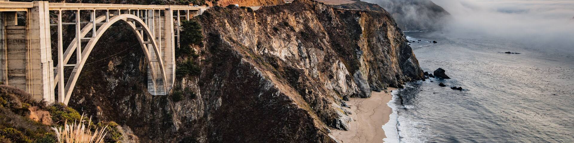 A bridge over a coastal cliff, ocean waves, and a mountainous landscape.