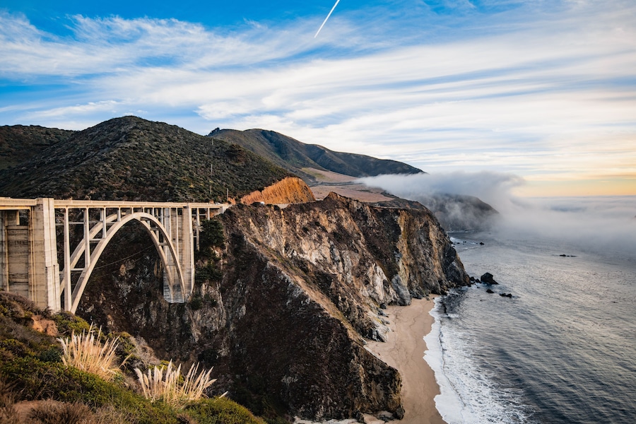 A bridge over a coastal cliff, ocean waves, and a mountainous landscape.