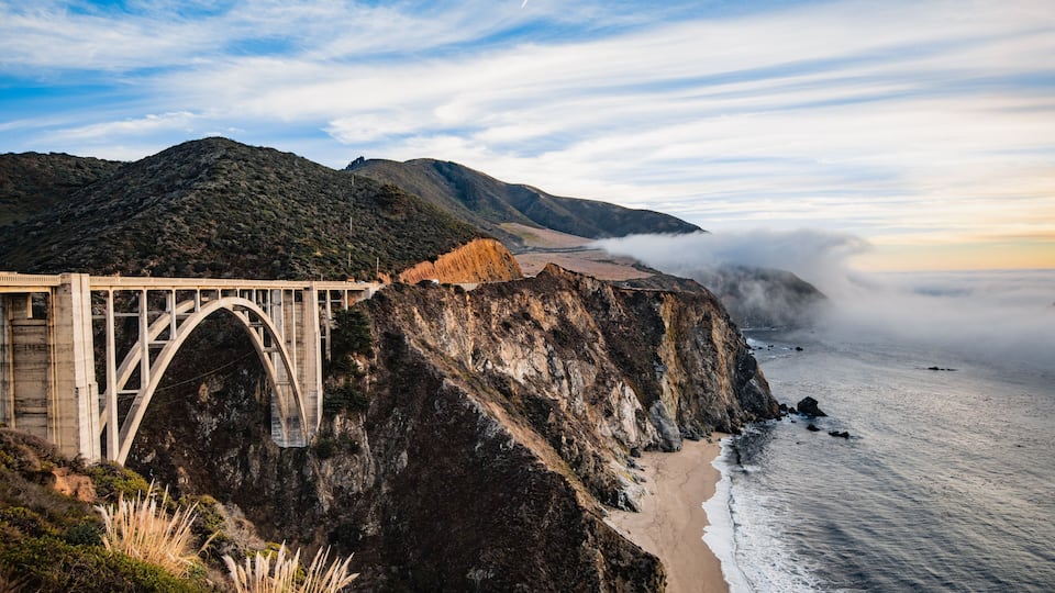A bridge over a coastal cliff, ocean waves, and a mountainous landscape.
