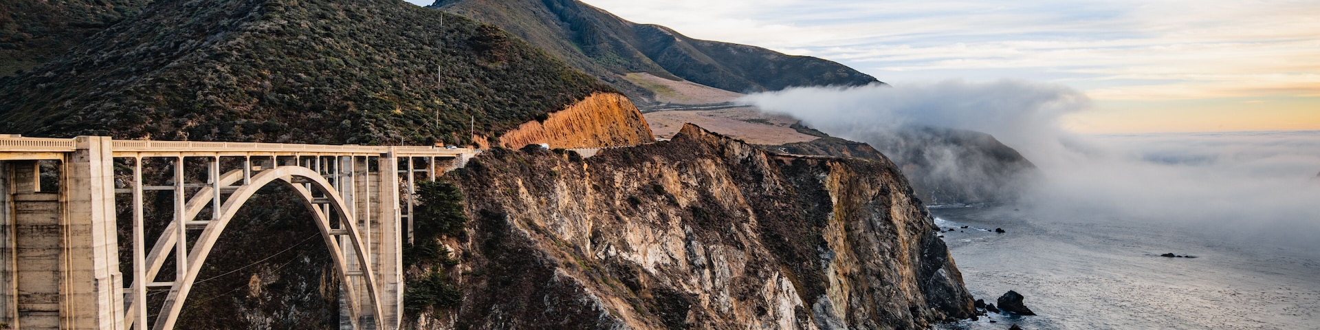 A bridge over a coastal cliff, ocean waves, and a mountainous landscape.