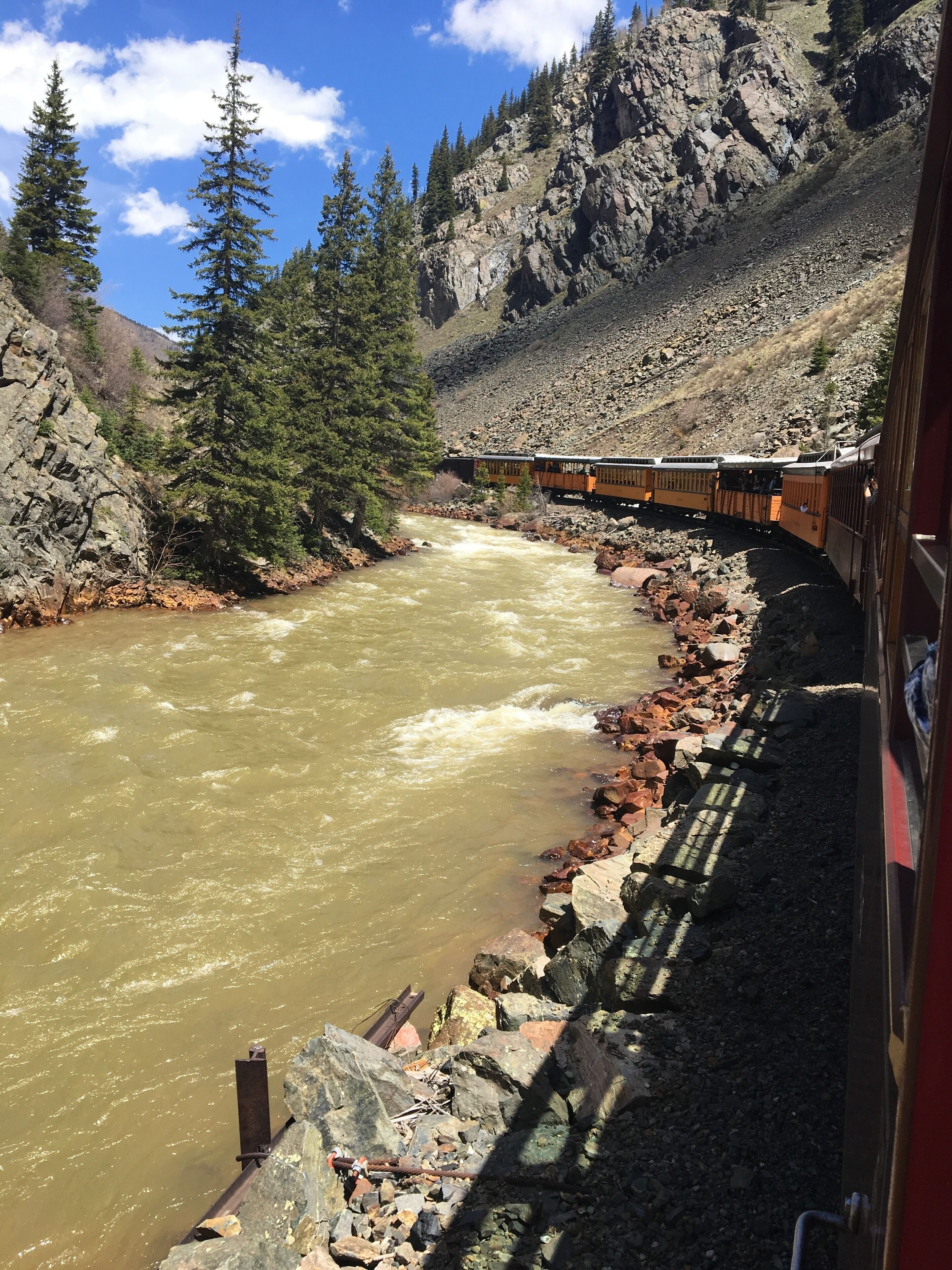 A train traveling along a rocky riverbed with a mountainous backdrop.