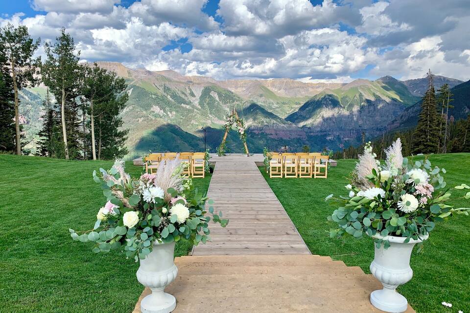 Un pasillo nupcial con sillas de madera y arreglos florales, frente a un paisaje montañoso.