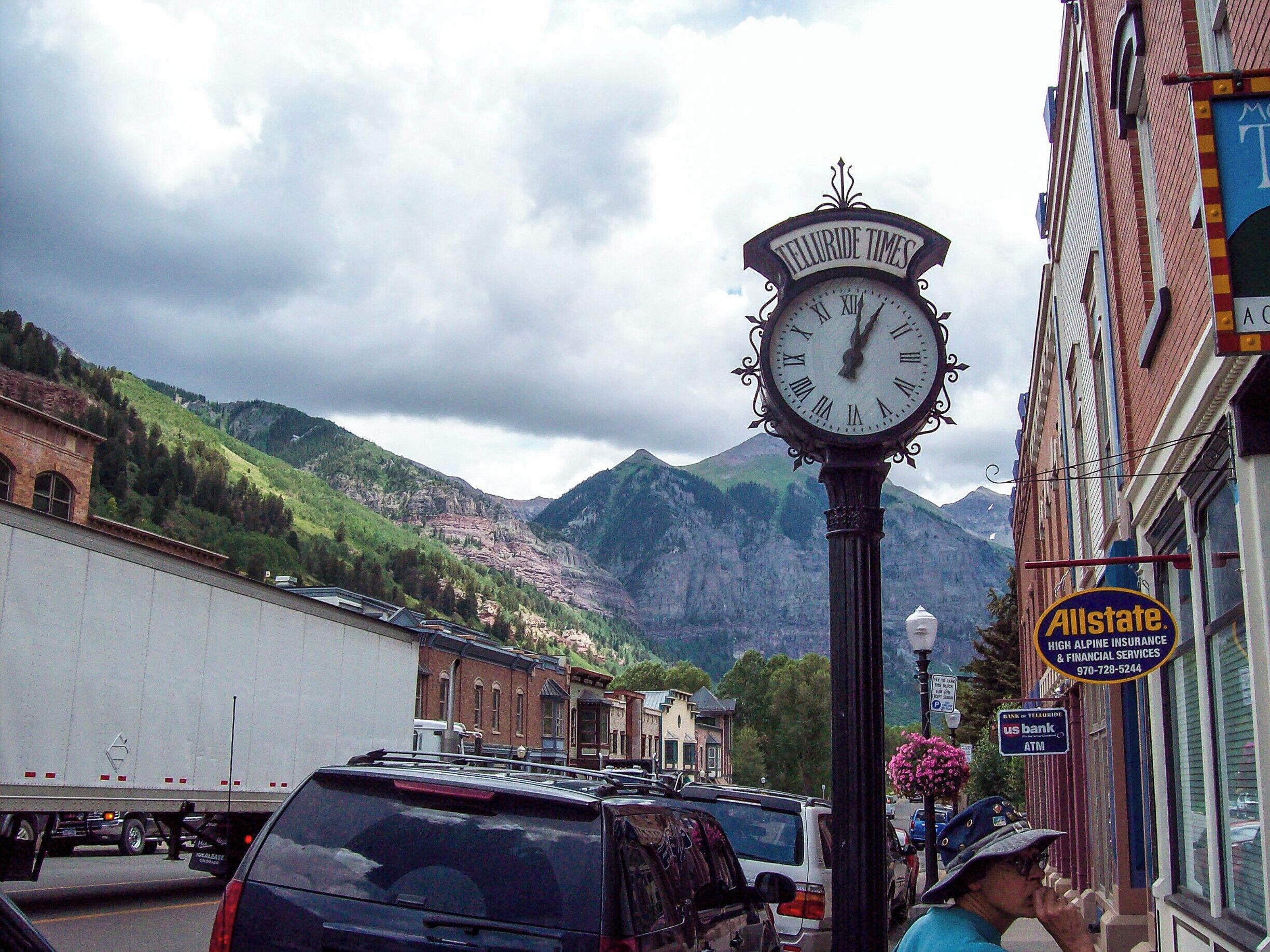 A clock with 'Telluride Times' on it, a mountain in the background, and a street with cars and buildings.