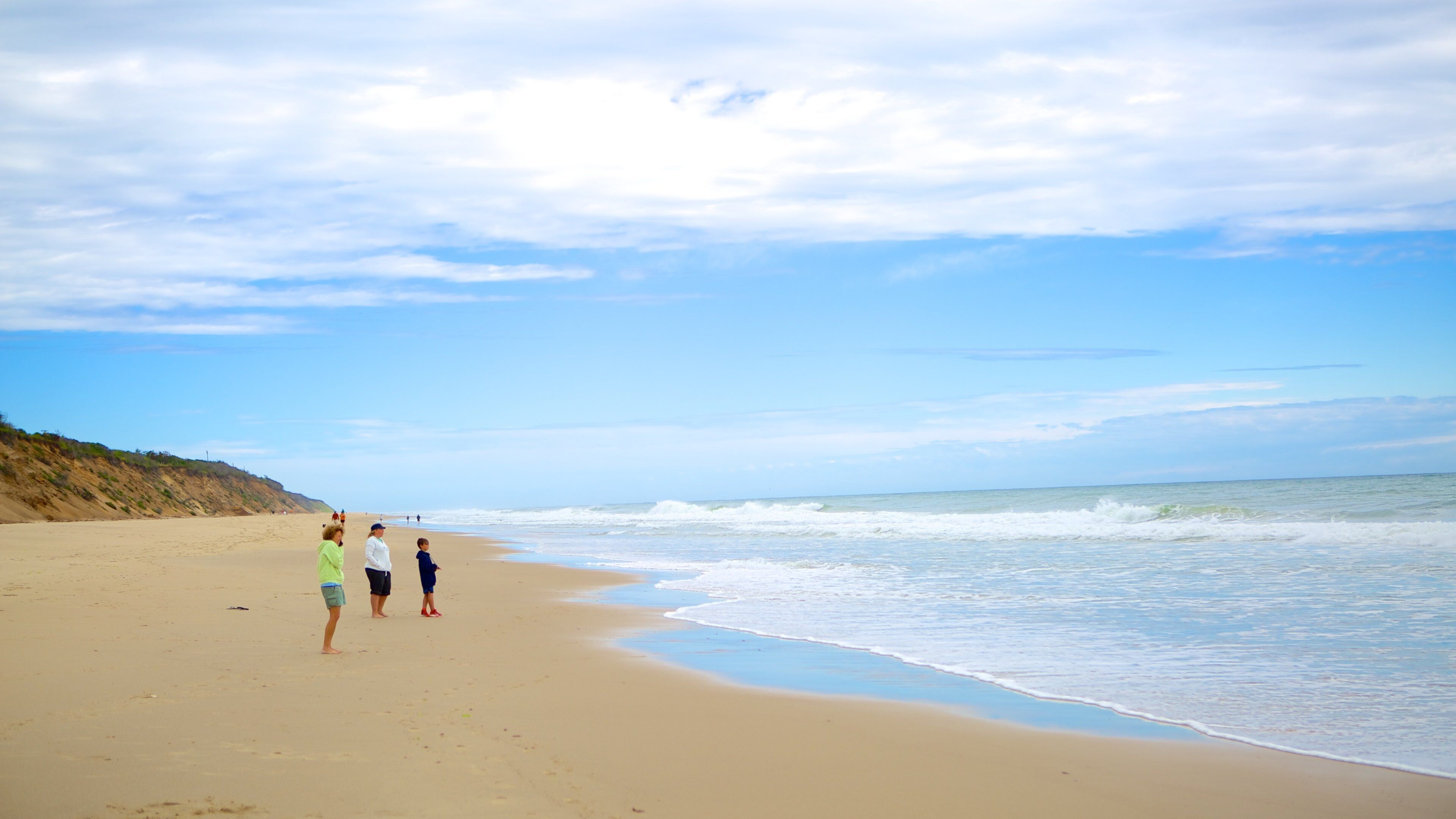 A sandy beach with gentle waves, a clear sky, and a distant cliff.