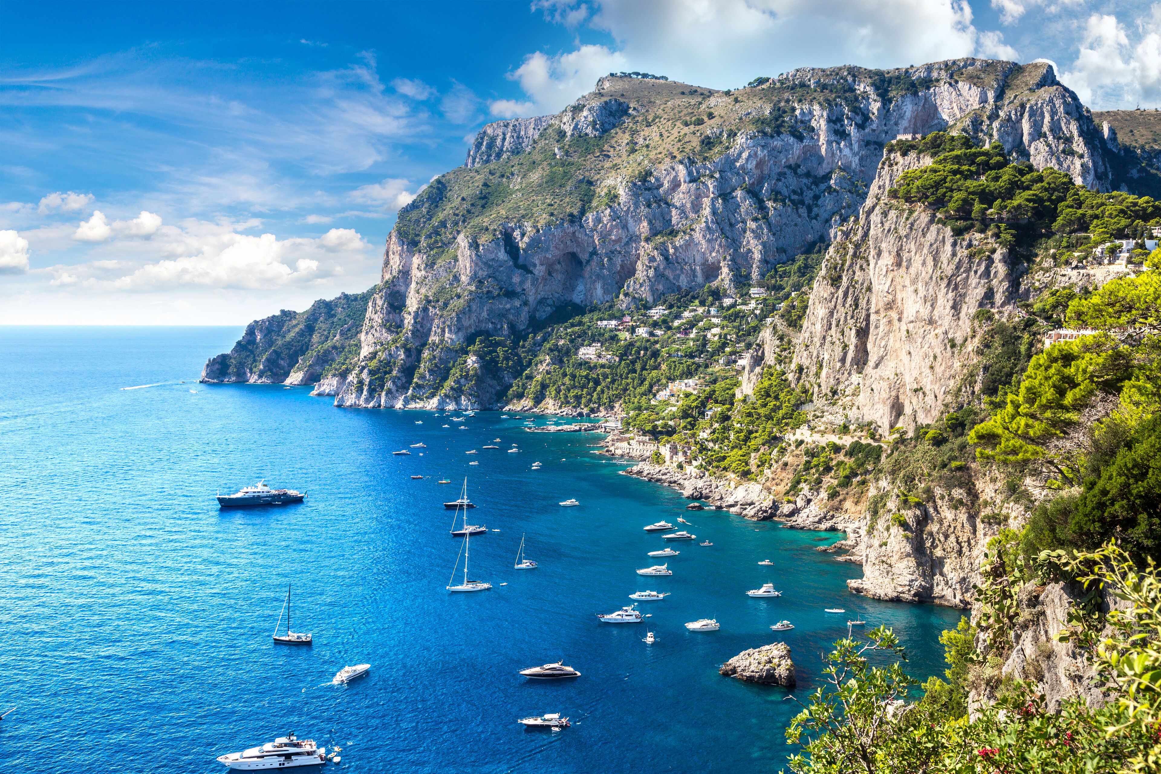 A coastal landscape with a rocky cliff, clear blue sea, and numerous boats anchored near the shore.