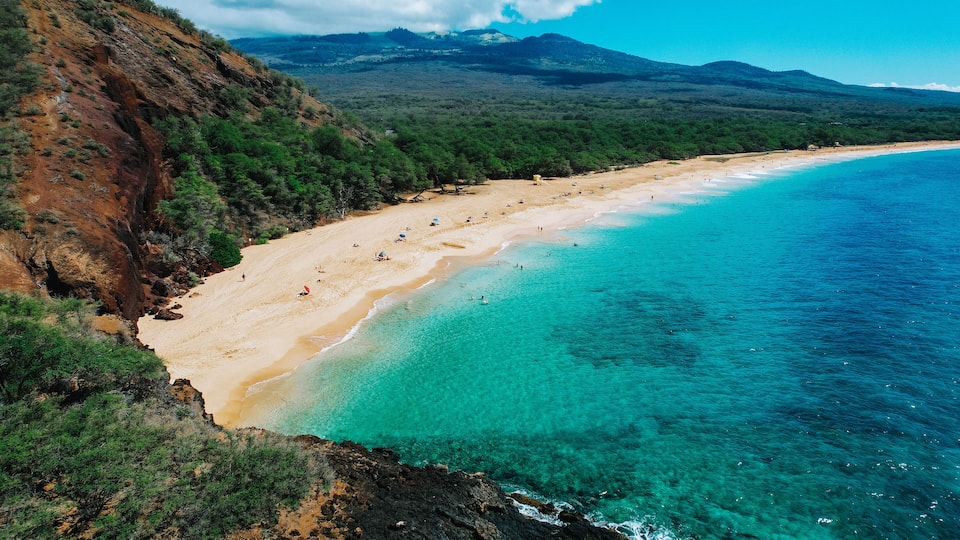 A coastal landscape with a sandy beach, clear turquoise waters, and a mountainous backdrop.