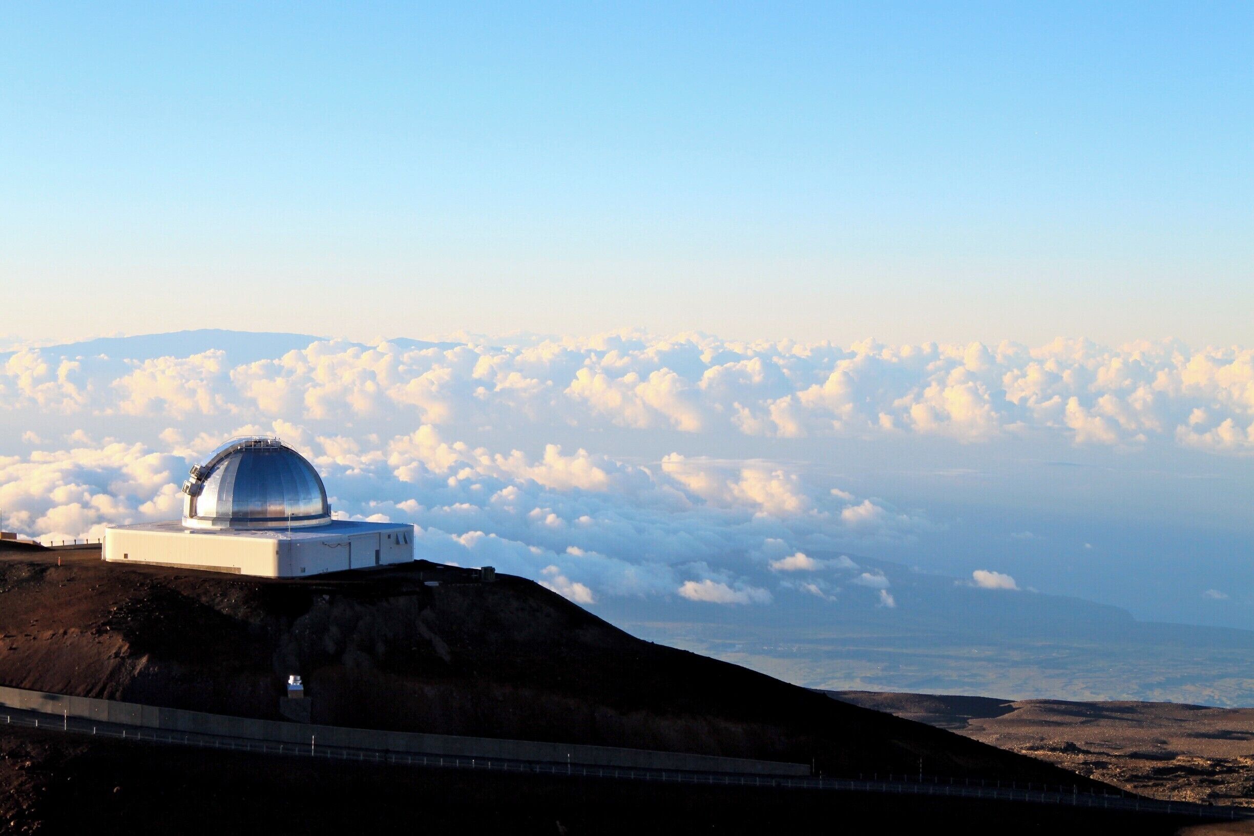 A dome observatory on a hill with a view of clouds below.