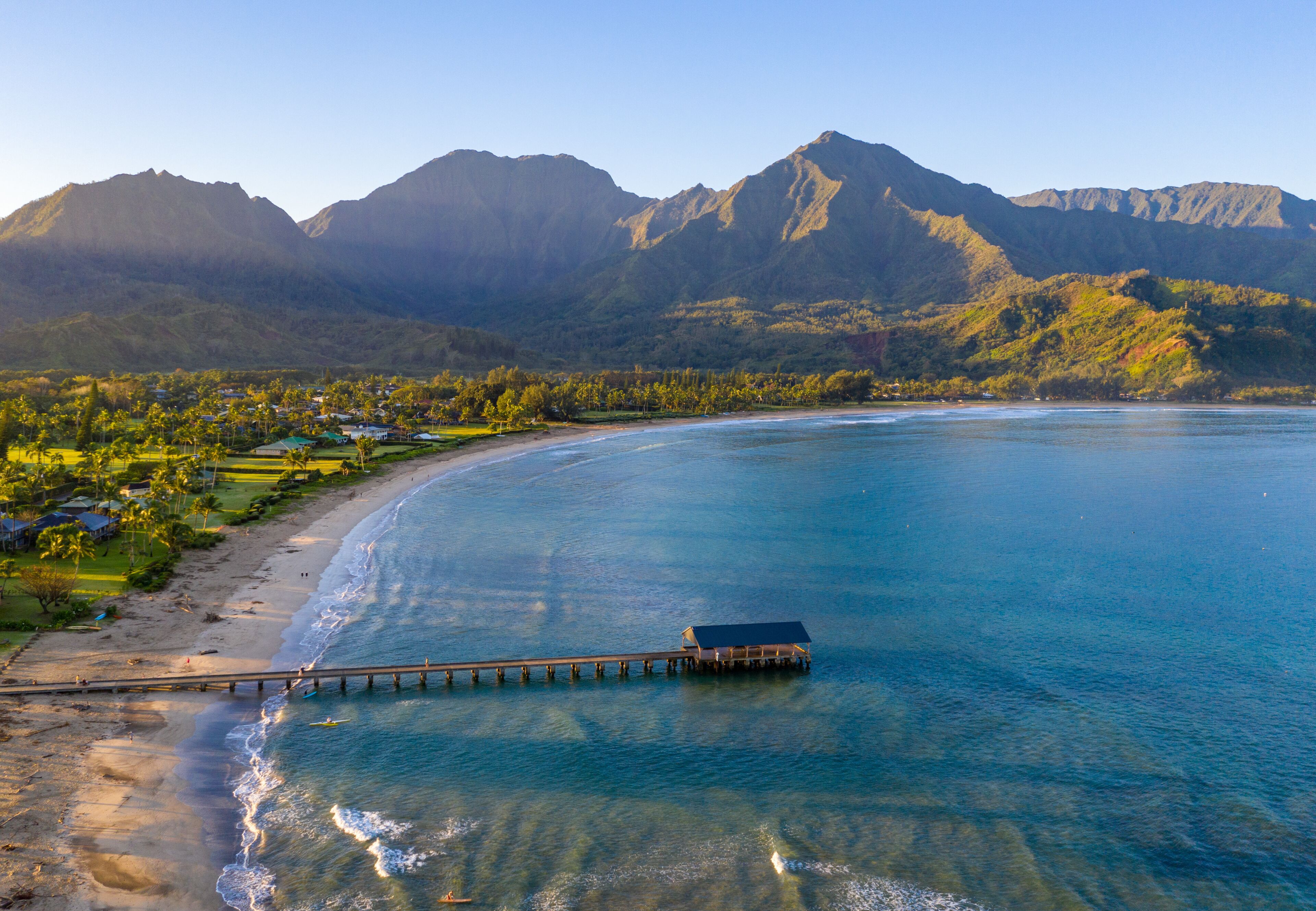 A coastal landscape with a pier extending into the ocean, surrounded by mountains and a residential area.