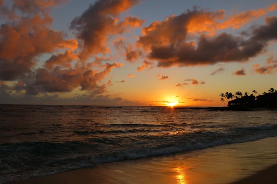 A sunset over the ocean with clouds and palm trees on the shore.