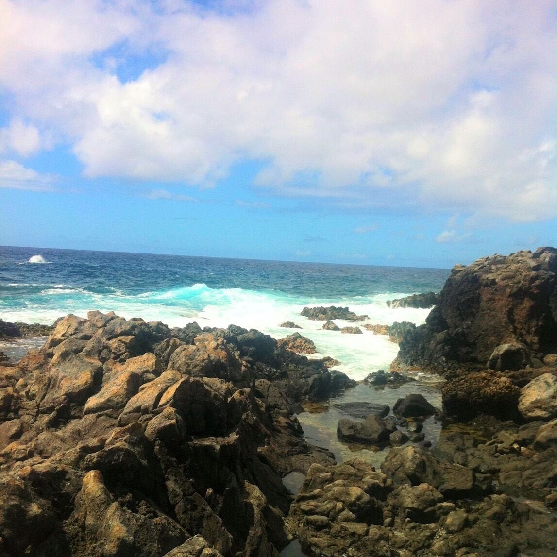 A rocky coastline with waves crashing against the rocks under a partly cloudy sky.