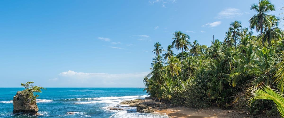 A tropical beach with clear blue waters, a rocky outcrop, and dense palm trees.
