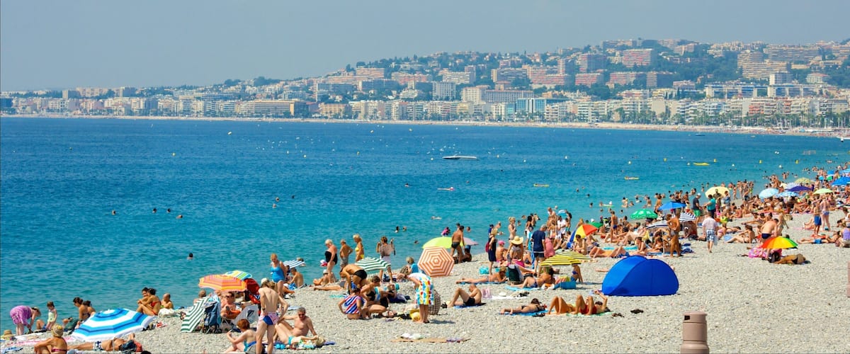 A crowded beach with people sunbathing and swimming, a cityscape in the background, and a clear blue sky.