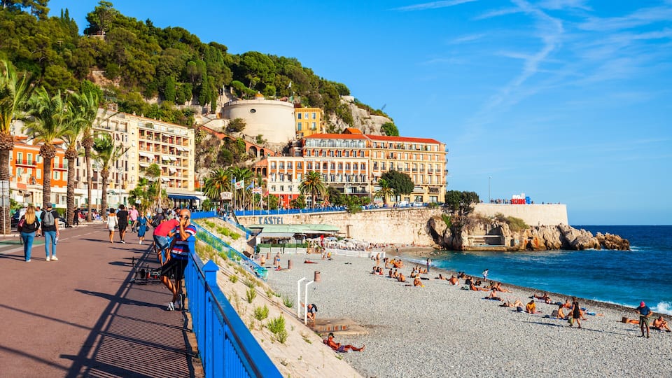A coastal promenade with a pebble beach, people sunbathing, and colorful buildings.