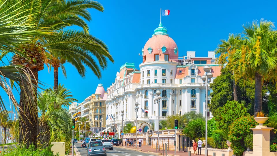 A grand building with a pink dome and a French flag, surrounded by palm trees and a clear blue sky.