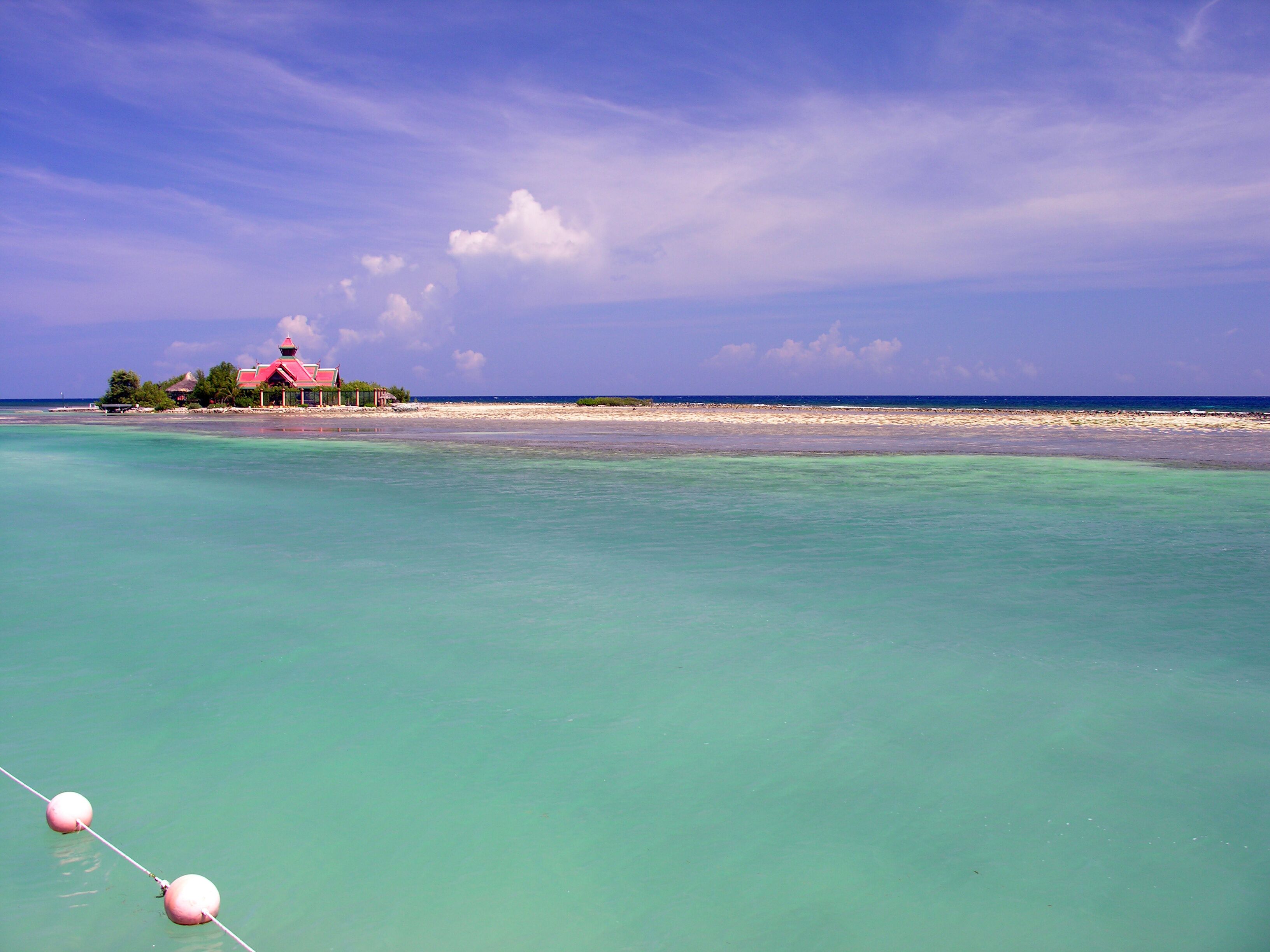 A beach with clear turquoise water, a small red-roofed building, and a distant shoreline.