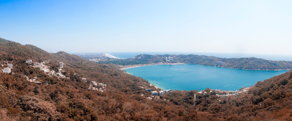 A coastal landscape with a bay surrounded by hills, buildings, and dense forests.