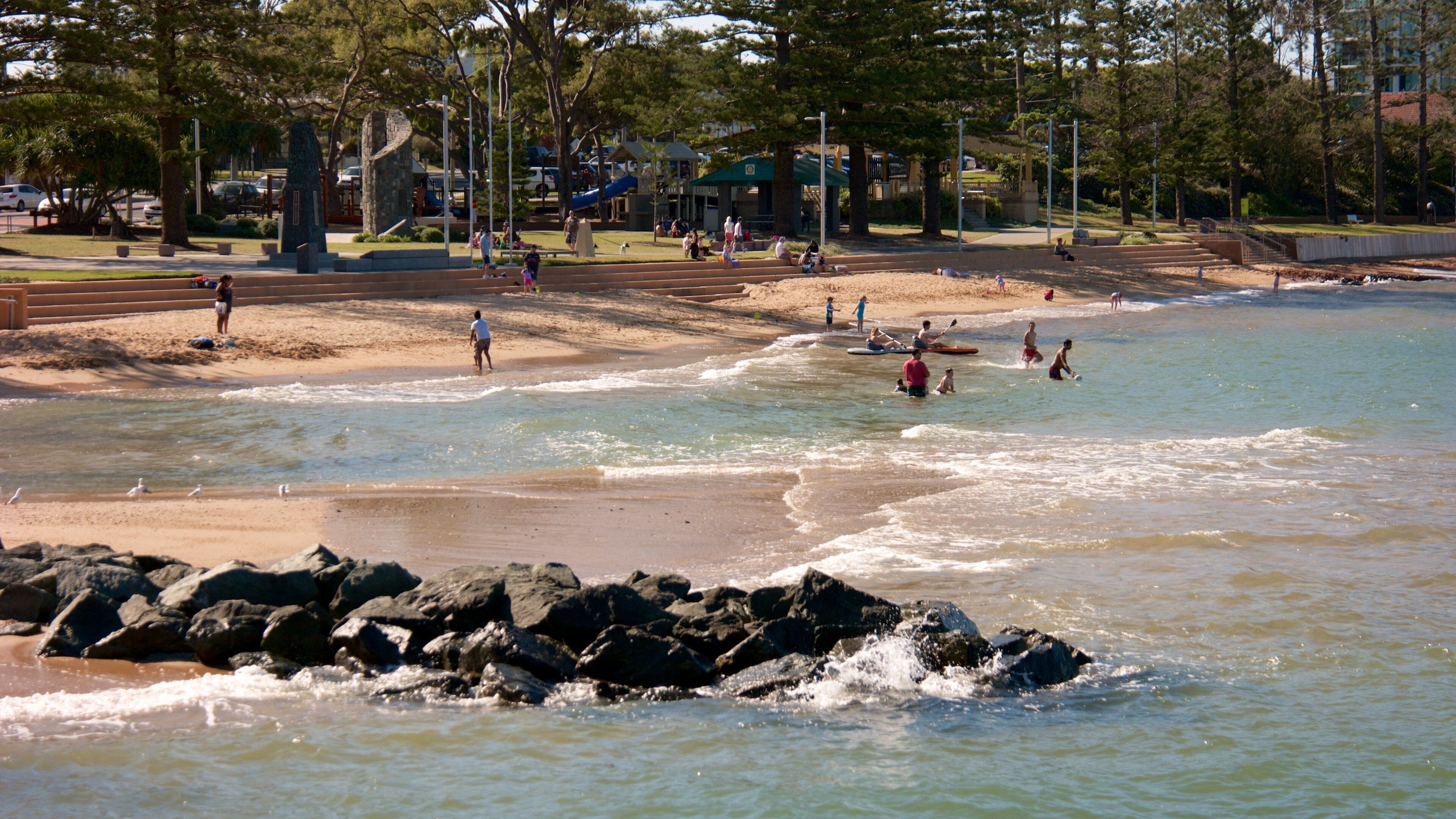 A beach with people swimming and walking along the shore, surrounded by trees and a park area.