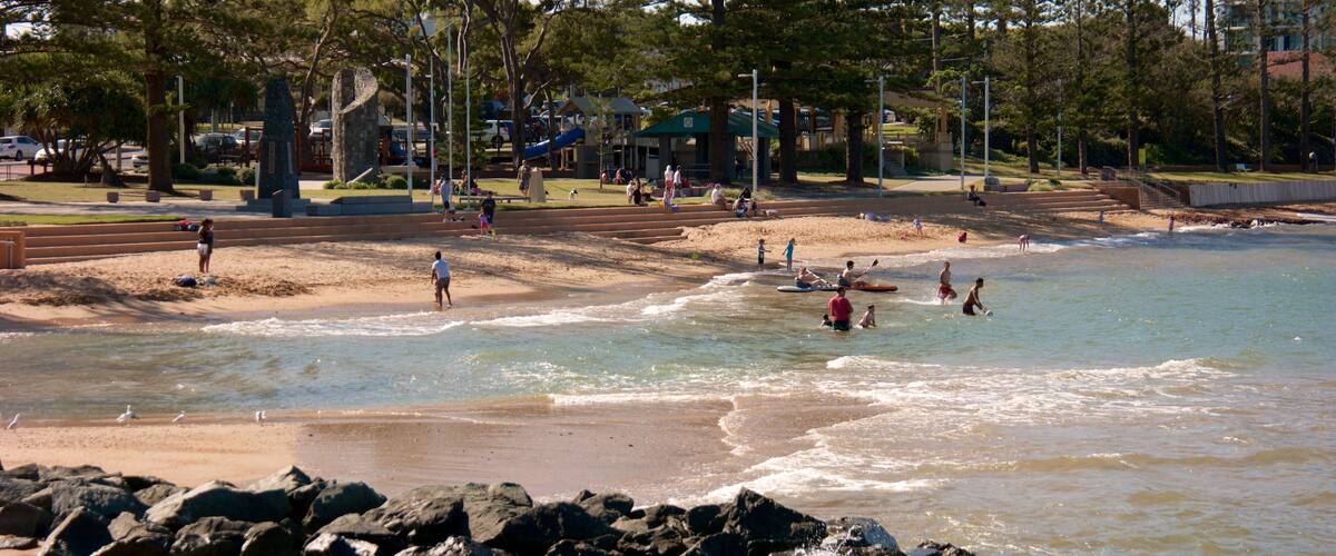 A beach with people swimming and walking along the shore, surrounded by trees and a park area.