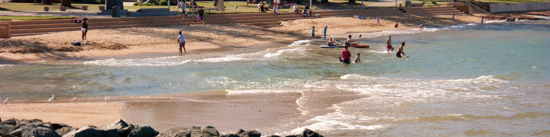 A beach with people swimming and walking along the shore, surrounded by trees and a park area.