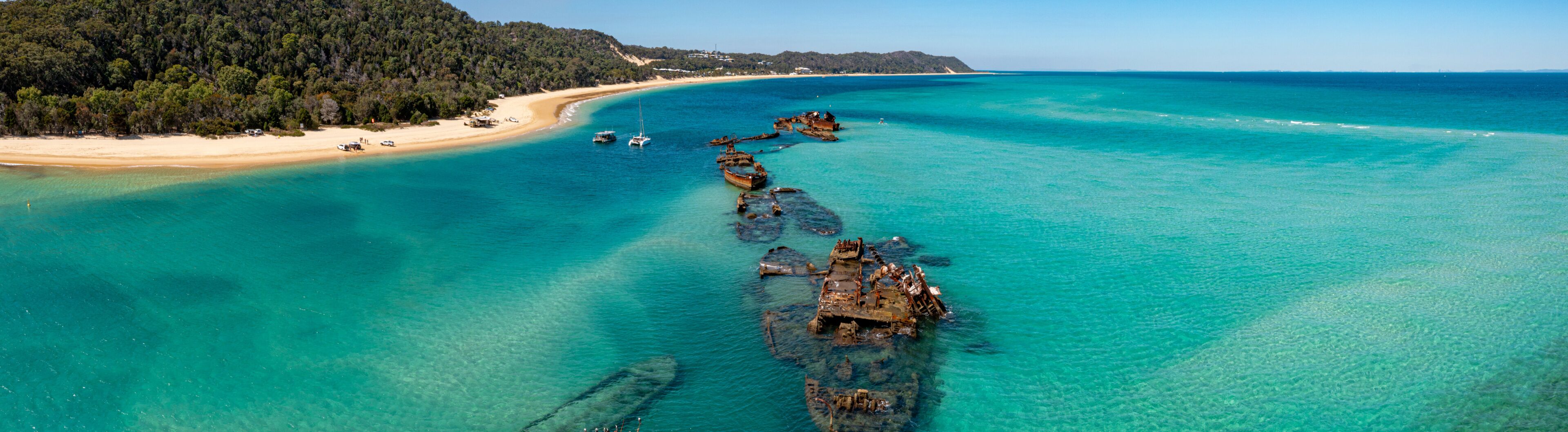 A coastal landscape with a sandy beach, clear turquoise waters, and submerged shipwrecks.