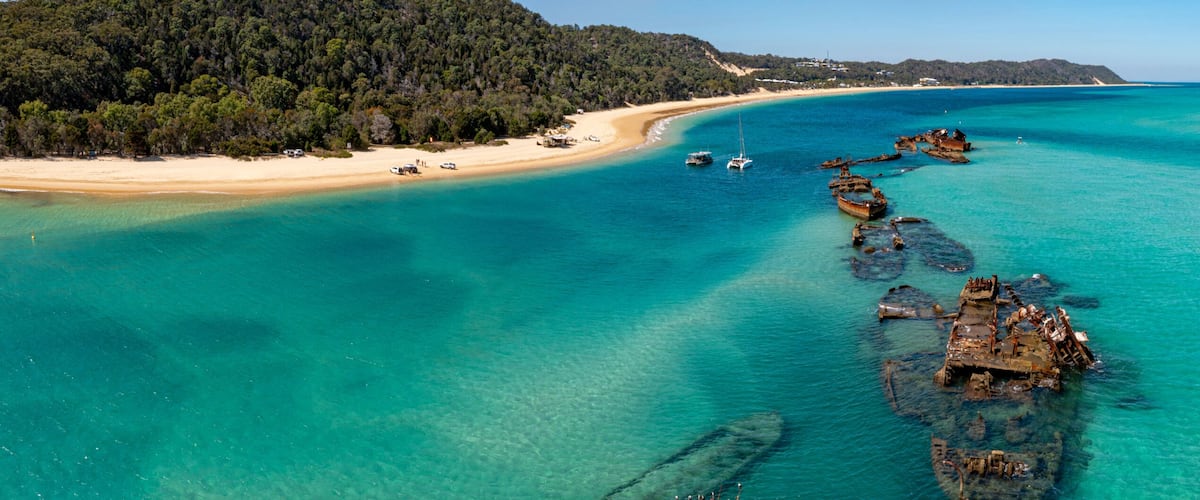 A coastal landscape with a sandy beach, clear turquoise waters, and submerged shipwrecks.