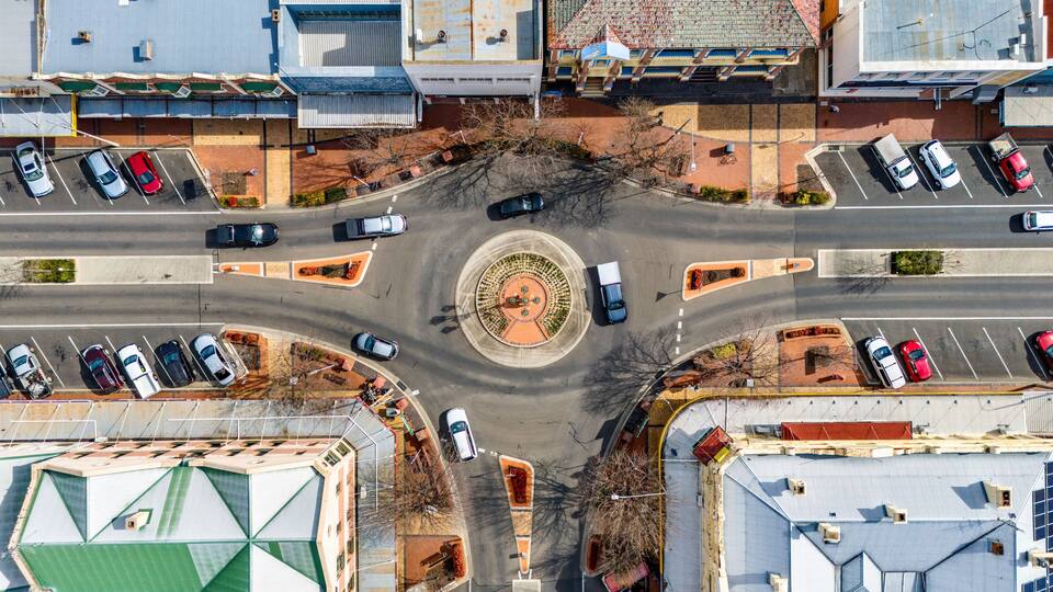 An aerial view of a roundabout with multiple lanes of traffic, surrounded by buildings and parked cars.