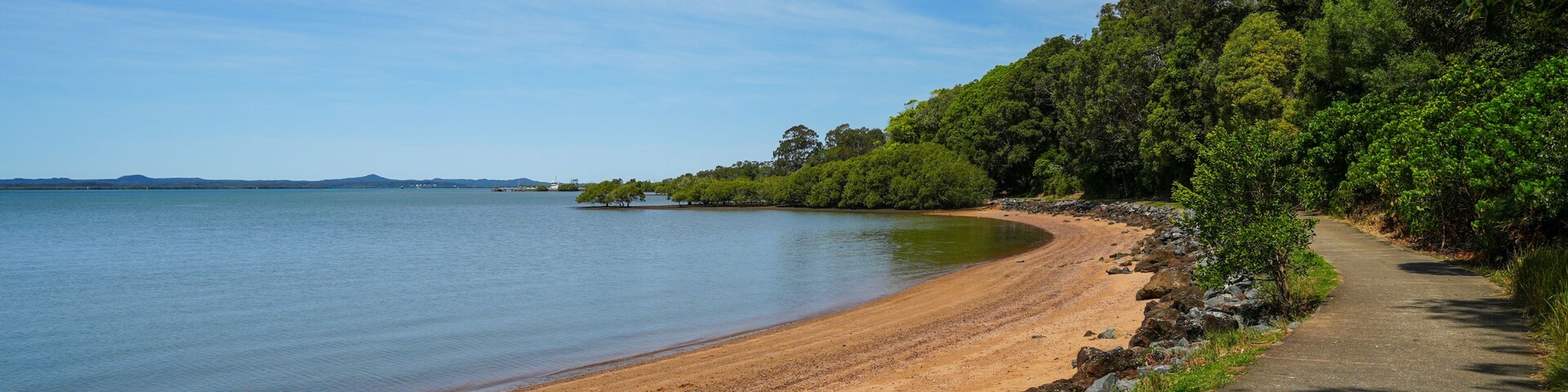 Una playa arenosa con un sendero curvo, agua tranquila y abundante vegetación verde.