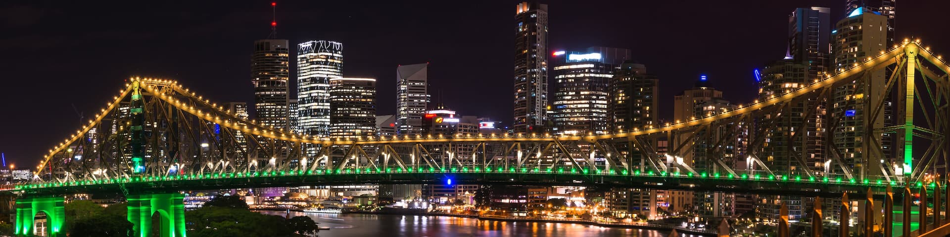 A brightly lit bridge with green lights spanning a body of water, surrounded by illuminated skyscrapers.