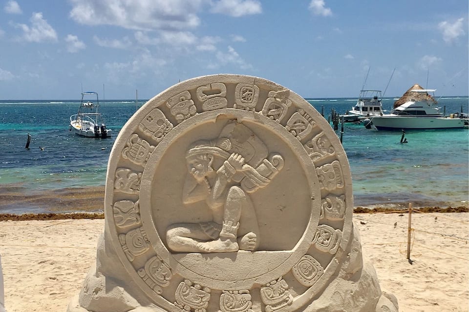 A carved stone sculpture on a sandy beach with boats in the background.