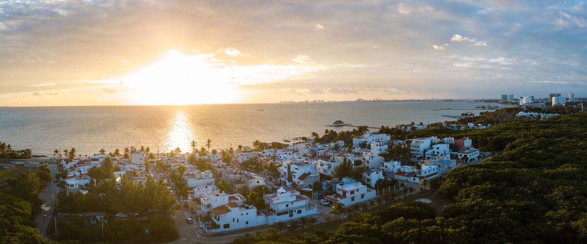 A coastal cityscape at sunset with white buildings, palm trees, and a distant city skyline.
