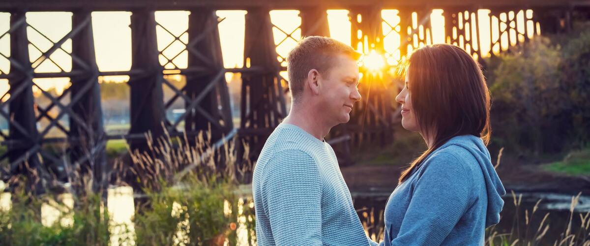 A couple embracing in front of a bridge with the sun setting in the background.