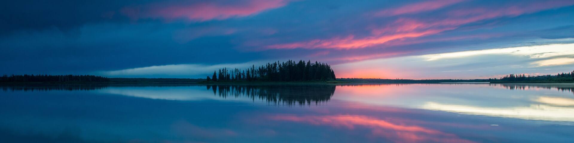A calm lake with a forested island and a colorful sky at dusk.