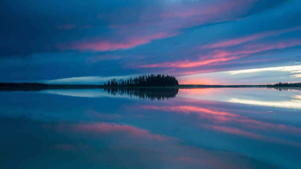 A calm lake with a forested island and a colorful sky at dusk.