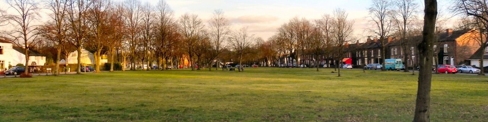 Un parque con un campo cubierto de césped, árboles sin hojas y una hilera de casas al fondo.