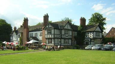 Um edifício em estilo Tudor com um gramado frontal, cercado por carros estacionados e pessoas.