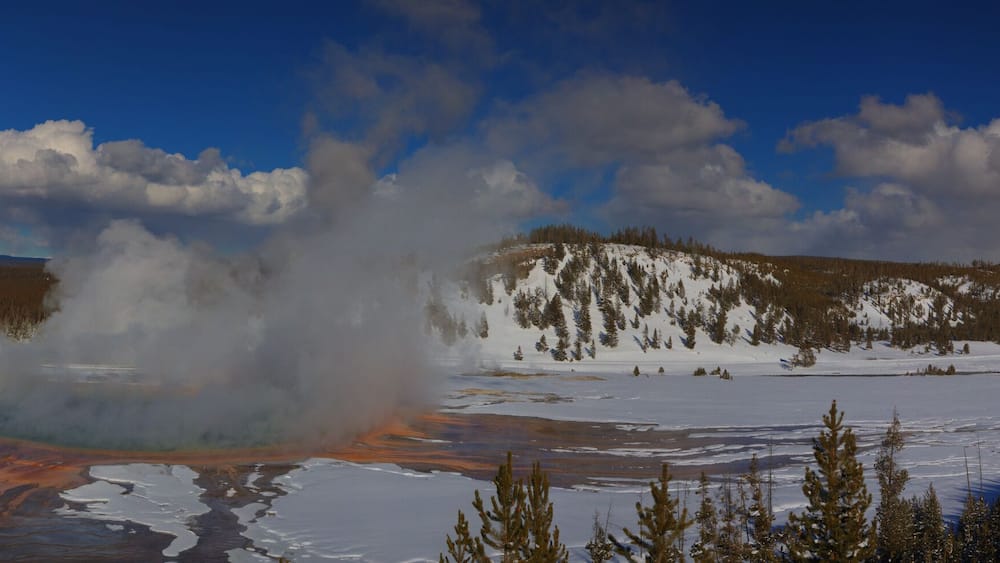 Ein Geysir bricht in einer verschneiten Landschaft aus, mit Bergen im Hintergrund und klarem Himmel.