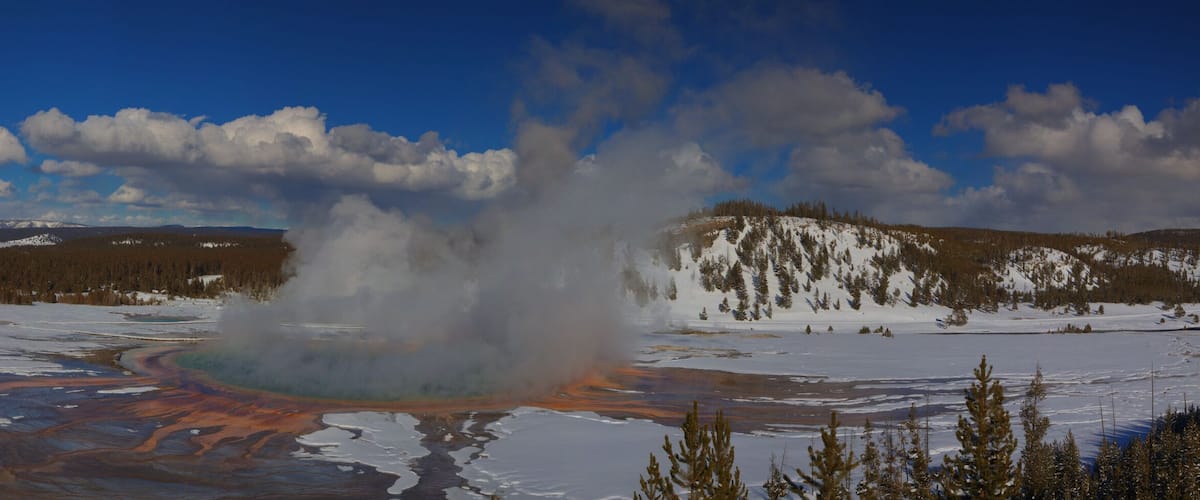 A geyser erupting in a snowy landscape with distant mountains and a clear sky.