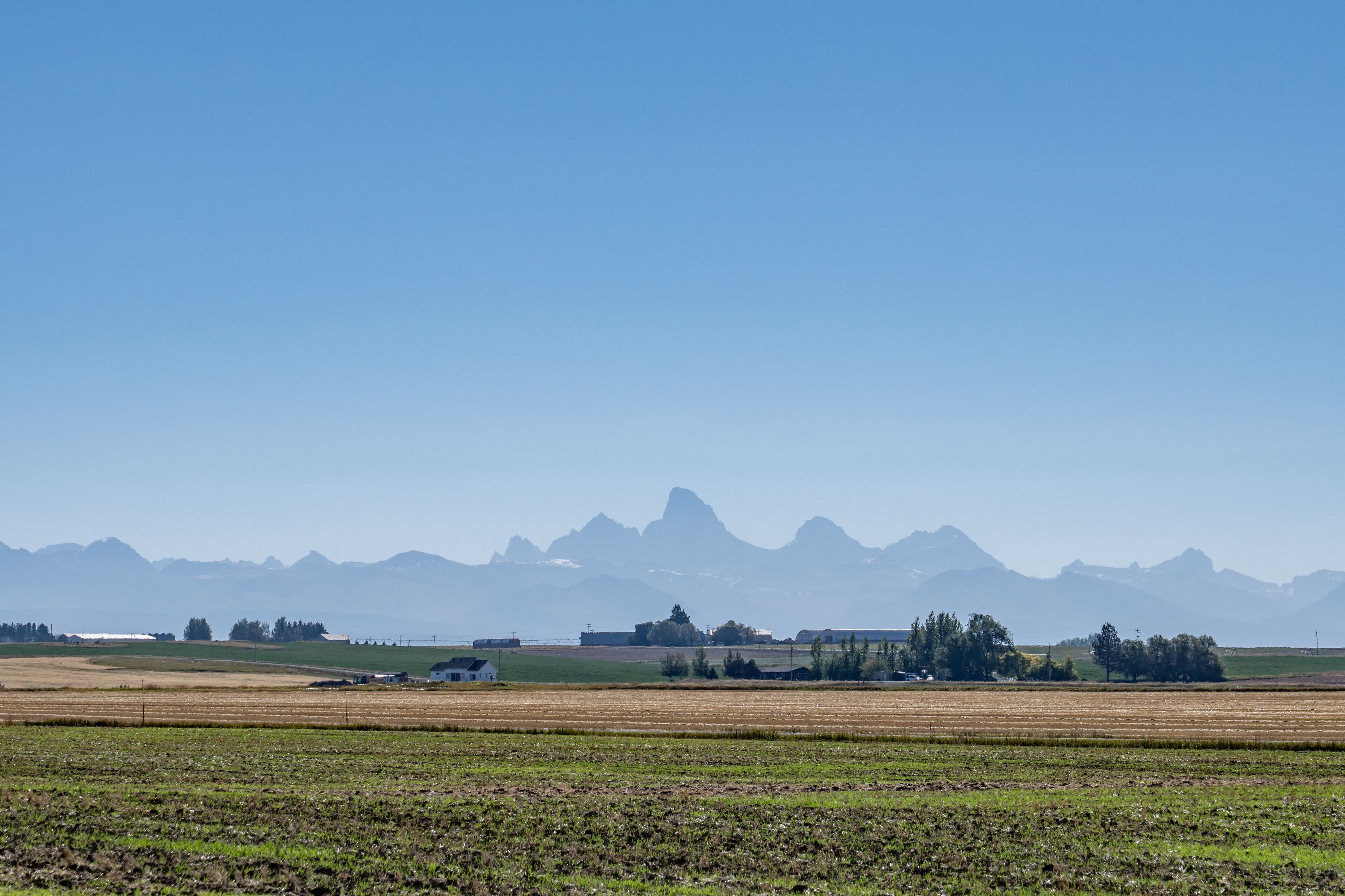 A rural landscape with fields, a small house, and distant mountains under a clear sky.