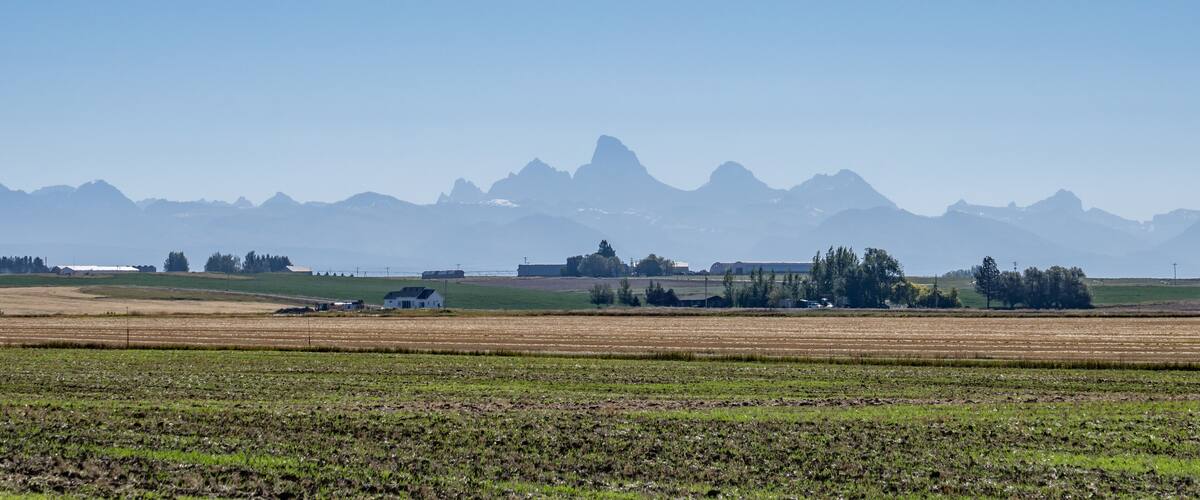 A rural landscape with fields, a small house, and distant mountains under a clear sky.
