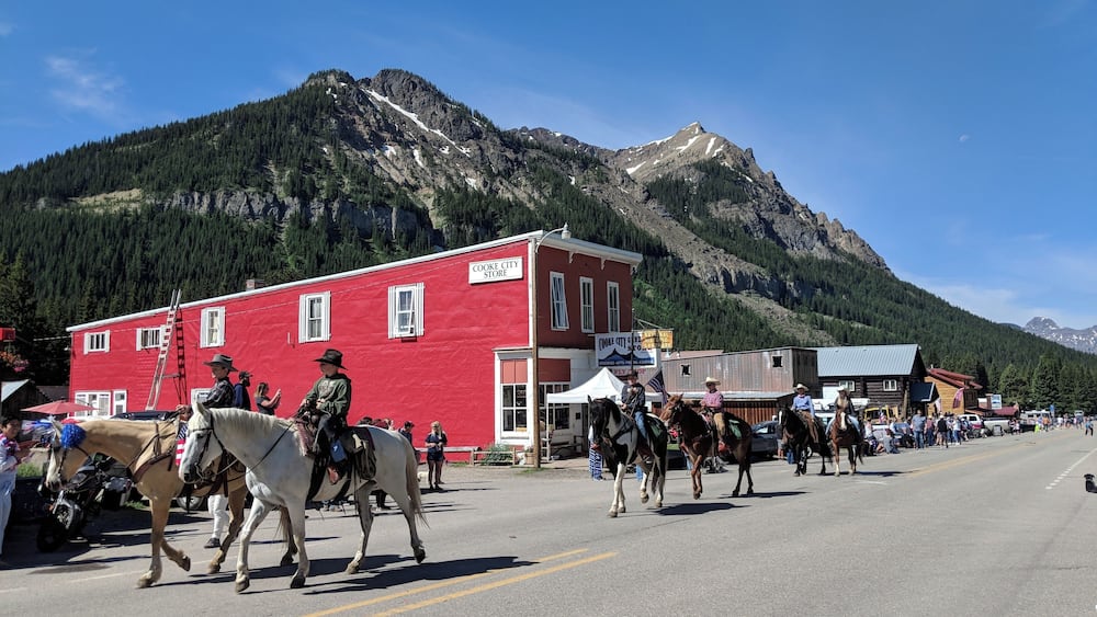 A street parade with people on horseback, a red building labeled 'COOKE CITY STORE', and a mountainous backdrop.