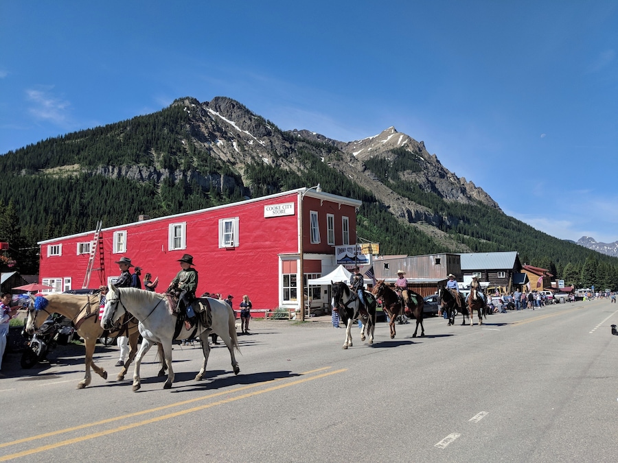 Un desfile callejero con gente a caballo, un edificio rojo con la inscripción 'COOKE CITY STORE' y un telón de montañas al fondo.