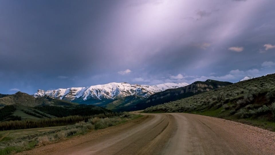 A dirt road curving through a mountainous landscape with snow-capped peaks in the background.