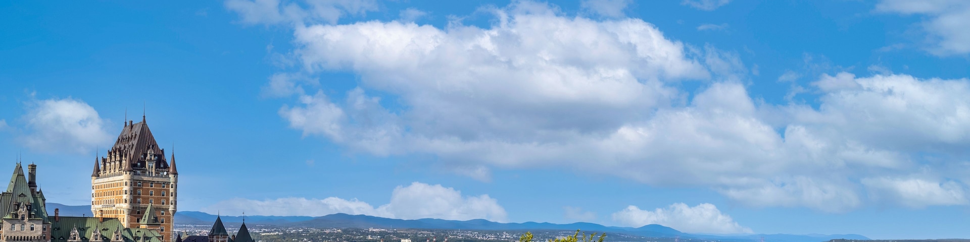 Un paisaje urbano costero con un castillo destacado, un río y montañas distantes bajo un cielo parcialmente nublado.